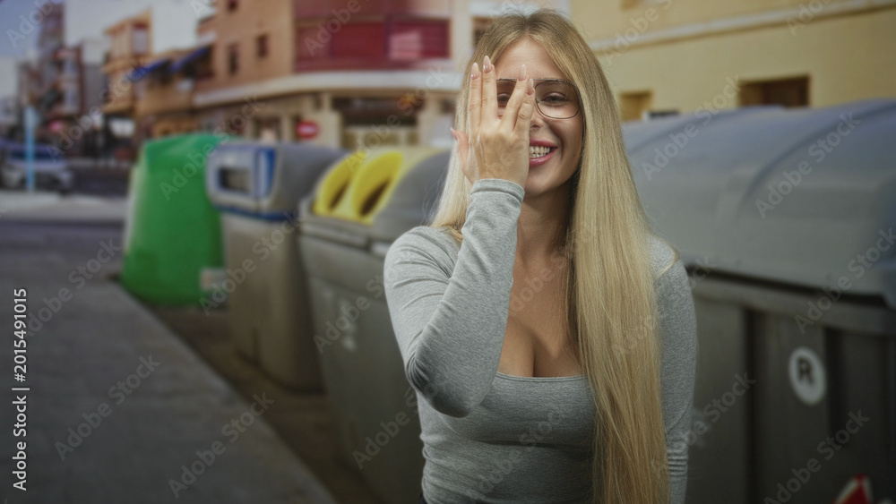 Fototapeta premium Woman covers eye with hand beside recycling bins on a street, wearing glasses with long blonde hair and smiling; playful joy.