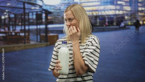 Quadro em tela Woman in striped shirt holding a milk bottle and biting fingernails on a street outside a glass building near a public plaza; anxiety hesitation uncertainty