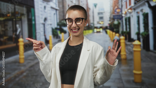 Bild auf Leinwand Woman pointing finger and showing ok hand on a cobblestone street, smiling with glasses and white blazer beside yellow bollards and shopfronts; playful confidence