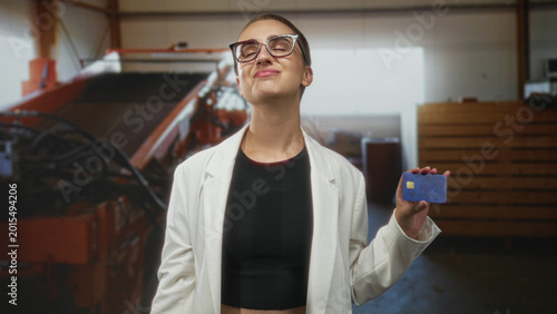 Photos Woman holding creditcard and smiling, shows the card with an outstretched hand while wearing glasses and a white jacket in building interior; trust confidence