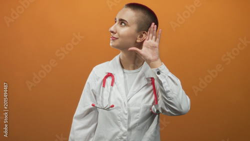 Foto Young woman doctor in white coat with red stethoscope cups hand to ear and tilts head while listening in studio; attentive care