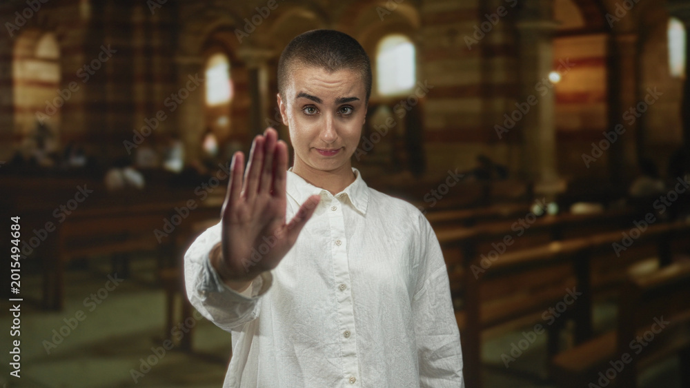 Naklejka premium Woman with short hair and white shirt holds hand raised palm forward in a building, making a stop gesture toward viewer; boundary setting defiance.