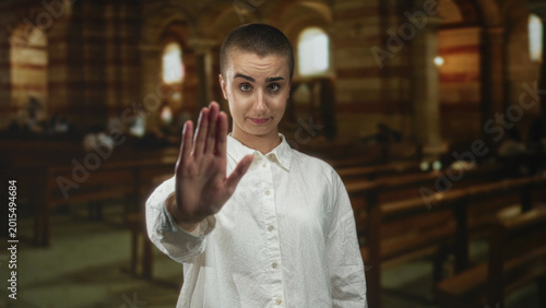 Vászonkép Woman with short hair and white shirt holds hand raised palm forward in a building, making a stop gesture toward viewer; boundary setting defiance