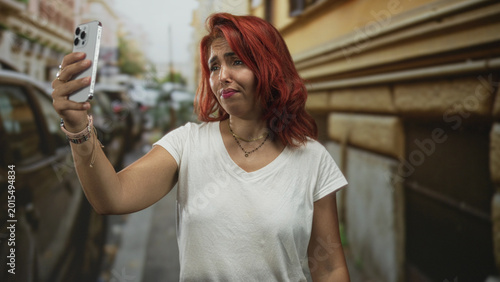 Φωτογραφία Woman holds smartphone in selfie pose on a city street near parked cars with a pained squint and pursed lips; frustration