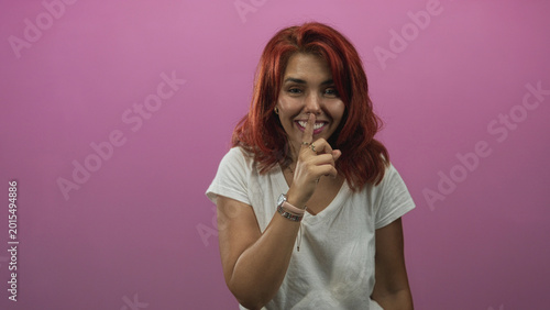 Obraz na plátne Woman with finger to lips for silence shushing toward camera in a pink studio wearing white t shirt and bracelets; playful secrecy