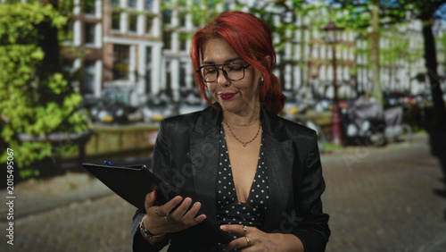 Canvastavla Red haired woman wearing glasses holds clipboard at cleavage level on a cobbled street in front of building; contemplative work focus