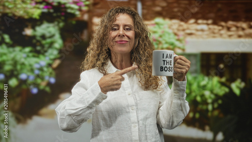 Photos Middle aged hispanic woman points finger to mug reading i am the boss in a building, smiling and holding cup in her right hand; confidence empowerment