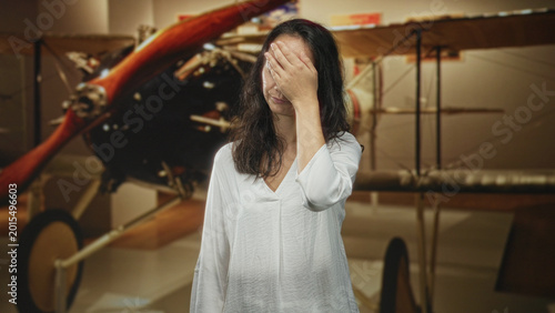Bild auf Leinwand Woman covers face with hand and makes expressive hand gestures beside a vintage biplane propeller inside building; frustration reflection