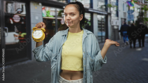 Fotografie Young hispanic woman holding yellow alarm clock and shrugging one hand in a busy city street wearing yellow crop top and denim shirt; frustration time pressure