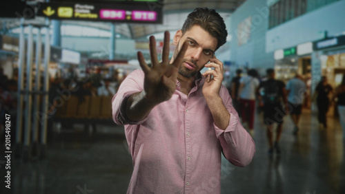 Konstfotografi Man holding smartphone to ear with hand palm out stop gesture at airport terminal crowd; stress urgency