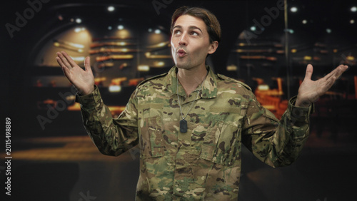 Tableau sur toile Man soldier in camouflage with dog tag and hands raised palms up, shrugging in a building beside rifle display under spotlights; pride duty