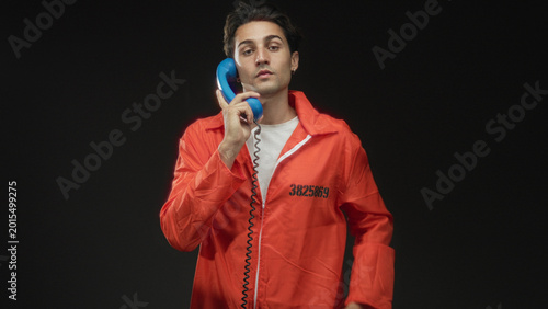 Quadro su tela Man inmate in orange jumpsuit holds a blue telephone receiver to cheek while folding arms in studio; melancholy solitude