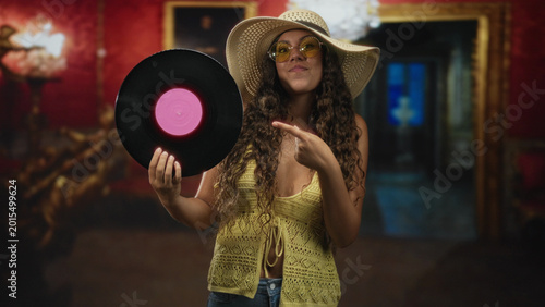 Slika na platnu Woman holding vinyl record with pink label and pointing finger at it while smiling in studio; playful retro nostalgia