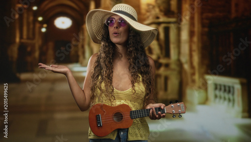 Foto Woman points finger and holds ukulele in an ornate building corridor wearing sun hat and round sunglasses while singing; playful travel curiosity