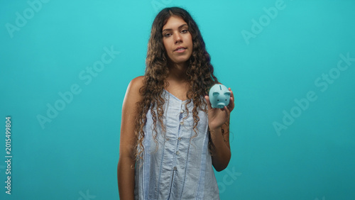 Fotografie Teen woman holding blue piggy bank in her hand and showing it to camera in a studio setting with turquoise backdrop; financial contemplation