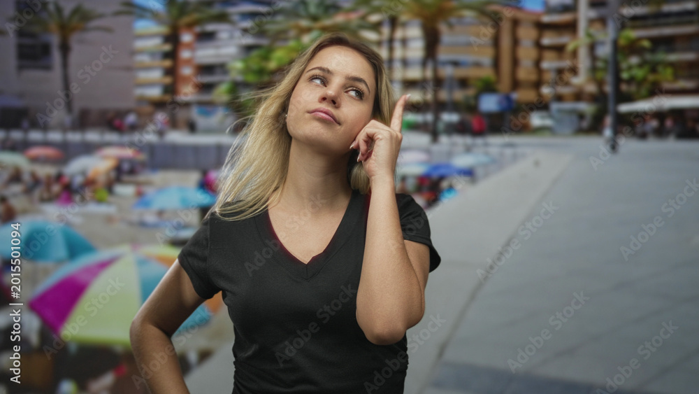 Fototapeta premium Young blonde woman points finger to temple while standing near a beachfront building in a vneck tshirt with hand on hip; thoughtful curiosity.
