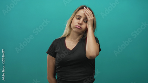 Tableau sur toile Young blonde woman wearing black t shirt, hand on forehead and eyes closed in a teal studio; stress fatigue