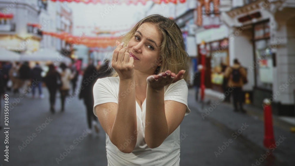 Fototapeta premium Young blonde woman with cupped hands offering to camera on a bustling city street, wearing white t shirt and looking forward; invitation warmth.