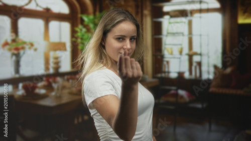 Fotografie Young blonde woman in white t shirt beckons with fingers in living room, looking directly at camera with a serious expression; challenge