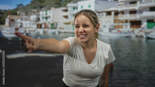 Tableau sur toile Young blonde woman points finger to nearby fishing boats beside a white coastal building at the port; anger accusation confrontation