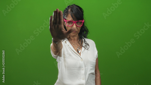 Foto Senior woman with pink glasses raises hand palm forward stop gesture in studio wearing white blouse and pendant necklace; personal defiance
