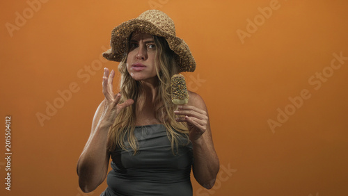 Konstfotografi Woman holding icecream bar in hand, straw hat and strapless grey dress in studio with orange backdrop; disgust hesitation