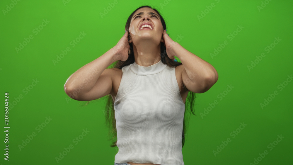 Fototapeta premium Woman with long dark hair in white tank top grimacing while covering ears in green studio; stress.