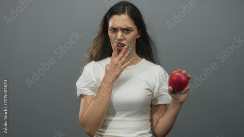 Canvastavla Woman with dark hair holds red apple with hand to mouth in gray studio setting wearing white shirt; disgust