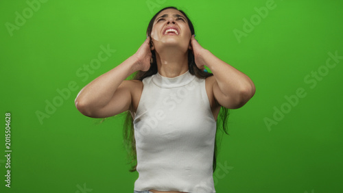 Tableau sur toile Woman with long dark hair in white tank top grimacing while covering ears in green studio; stress