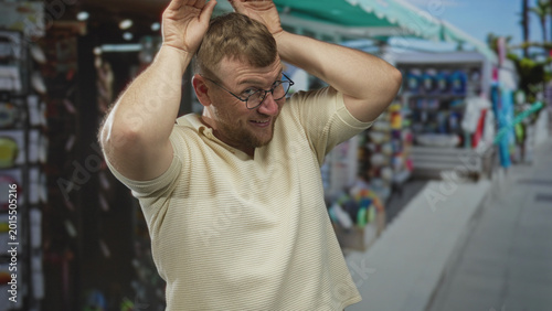 Fotografía Man with hands to head making antler gesture on a busy street near souvenir stalls, wearing glasses and short sleeve knit shirt; playful mischief