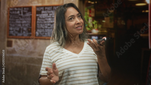 Bild auf Leinwand Hispanic woman holding smartphone and gesturing while speaking into phone in a building cafe interior with chalk menu and counter; frustration