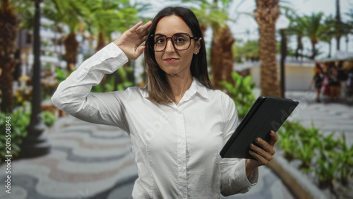 Fényképezés Woman in white shirt tapping a tablet with fingers while holding the folio cover on a palm lined pedestrian street with benches and plants; focused productivity