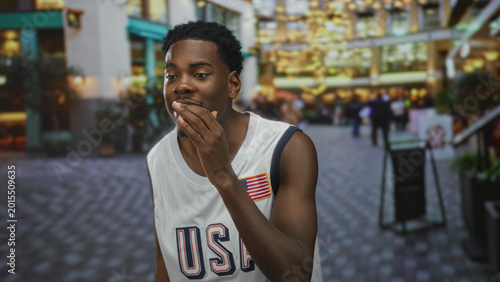 Quadro su tela Man wearing usa jersey waves hand along bustling city street lined with illuminated storefronts and buildings; eagerness connection