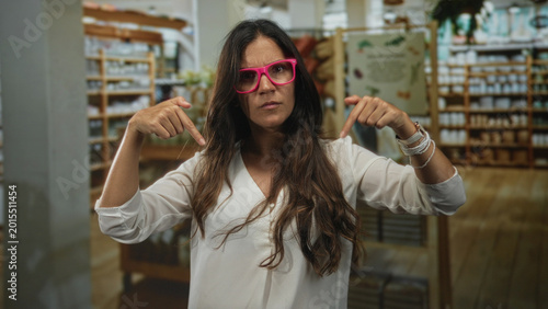 Cuadro en lienzo Woman wearing pink glasses pointing both index fingers downward in building aisle showing forearms and bracelets; assertive confidence
