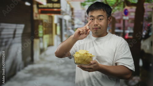 Photos Young chinese man holds a clear glass bowl of potato chips with both hands on street under midday sunlight; curiosity craving