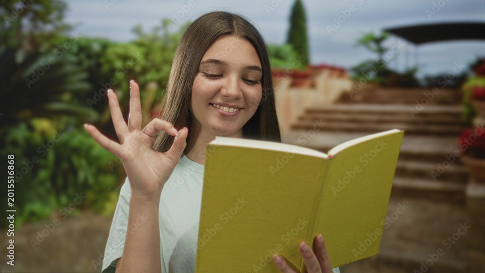 Fototapeta premium Teenage girl reading a green paperback book on stone steps in a park outdoors, holding the book; calm learning concentration.