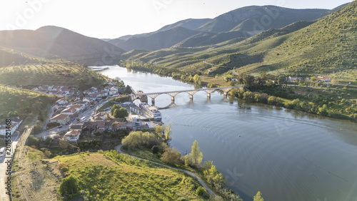 Aerial view of Barca de Alva village and the bridge over the Douro River, surrounded by terraced vineyards and green hills in Figueira de Castelo Rodrigo, Distrito da Guarda, Portugal.