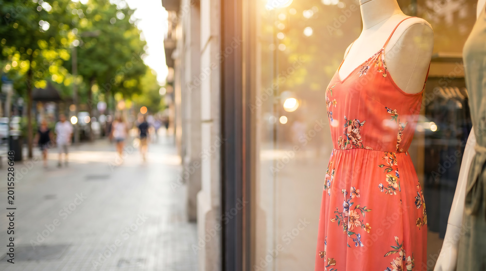 Obraz premium Female mannequin dressed in a floral summer dress displayed in a shop window, with a sunlit street and blurred pedestrians visible in the background