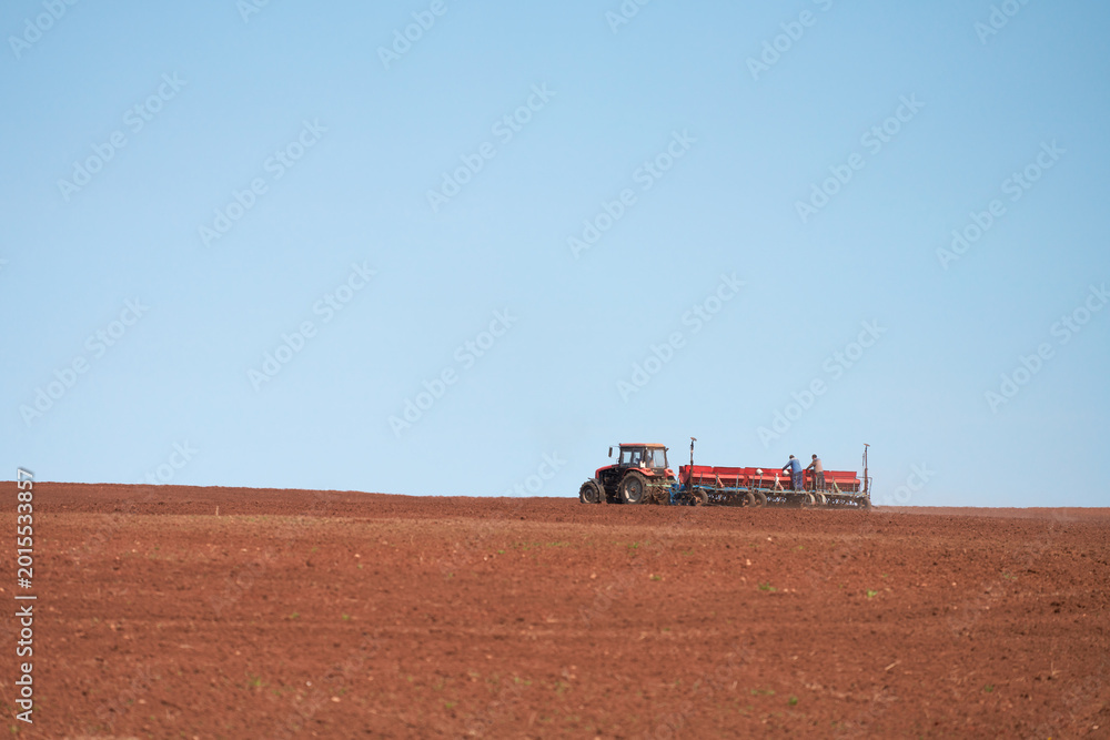 Fototapeta premium Spring sowing campaign. A tractor with a towed seeder is working in the field. Two workers are on the seeder, monitoring the quality of the seed placement.