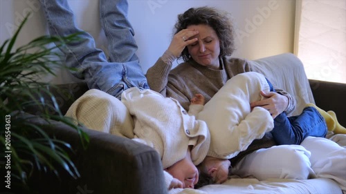 Exhausted mother experiencing a headache while her two hyperactive twin children are uncontrollably jumping upside down on the sofa in the living room, a challenging moment in motherhood