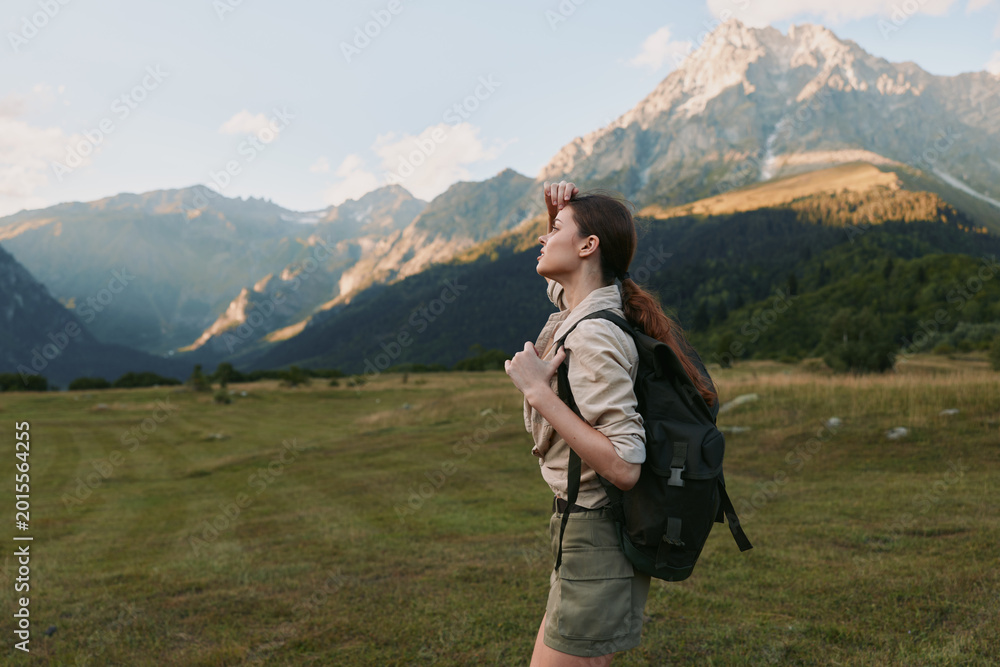 Fototapeta premium Young woman with a backpack hikes across a grassy valley toward distant mountains, captured in soft light that highlights exploration, independence and a deep connection with nature.