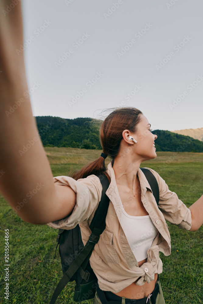 Naklejka premium A woman with a backpack raises her arms in a bright outdoor field, smiling, embracing nature and freedom during a hiking adventure.