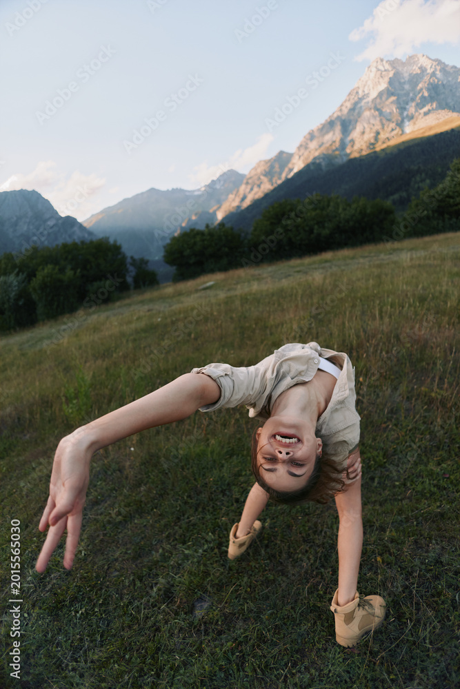 Naklejka premium Young woman performs a backbend in a grassy meadow with distant mountains, capturing a playful, adventurous moment outdoors.