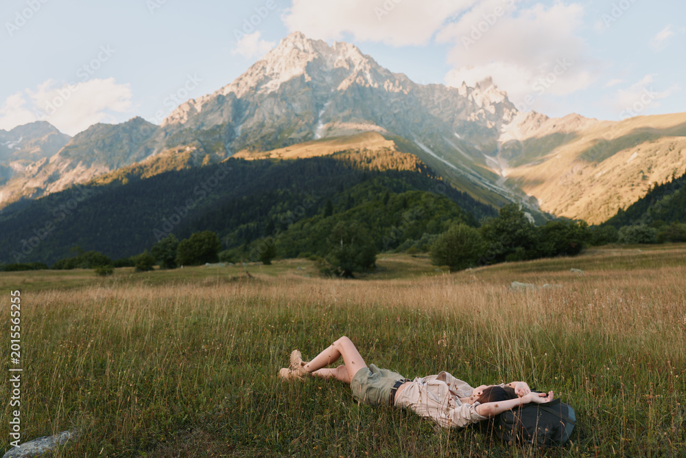 Obraz premium Young woman lies in a sunlit meadow with mountains in the background, relaxed pose and peaceful expression amid tall grass, a gentle breeze, warm light, natural colors, distant pines, and wide open