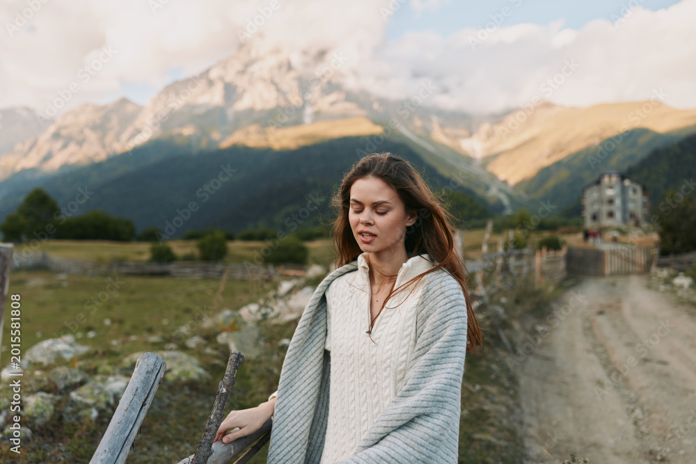 Obraz premium A young woman with brown hair walks along a rustic fence beside a mountain tapestry wrapped in a light knit sweater calm and thoughtful in the open countryside