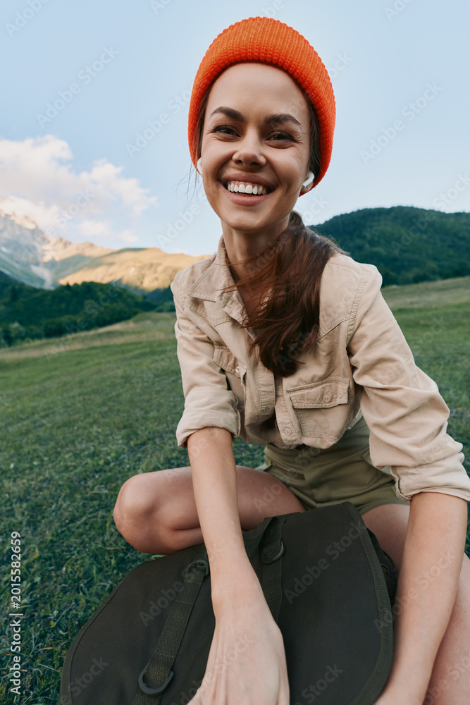 Naklejka premium Cheerful woman lifestyle outdoors, smiling on a sunlit mountainside field, casual beige shirt and orange beanie, capturing a carefree moment with nature and wide landscape