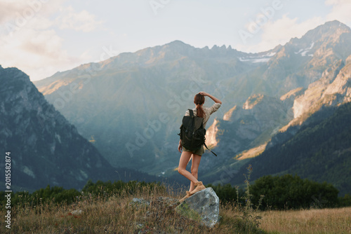 Fotografie Young woman with backpack stands on a rock in a mountain valley, looking toward