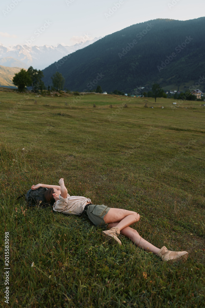 Obraz premium Relaxed woman lying on grass in a wide countryside field with distant mountains, capturing a casual outdoor lifestyle moment of rest, freedom, and connection with nature.