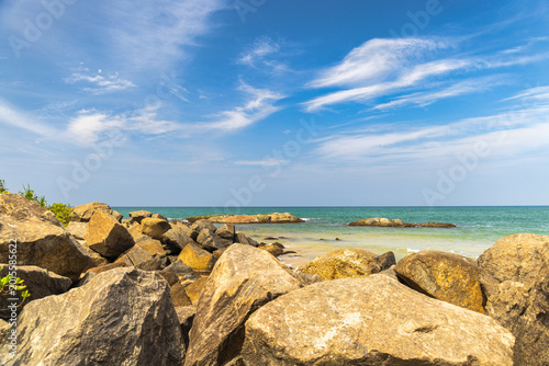 Rugged coastal landscape with large brown rocks bordering a serene turquoise ocean under a vibrant blue sky with wispy white clouds. Sri Lanka island in Asia.