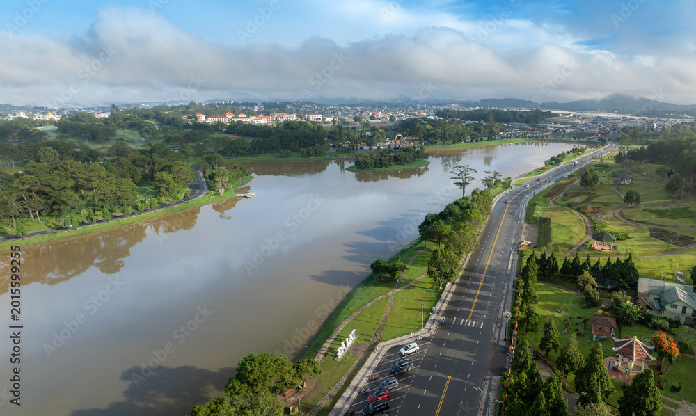 Fototapeta premium An aerial view of Xuan Huong Lake in Da Lat, Vietnam, on a cloudy morning. The lake is surrounded by pine trees and a city skyline.